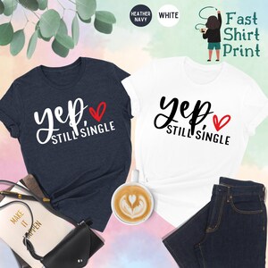 May include: Two t-shirts, one heather navy and one white, with the text "yep, still single" and a red heart. The shirts are laid out on a pink and blue background with a cup of coffee, a book, and a purse.