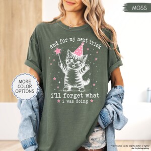 May include: A moss green short-sleeve t-shirt featuring a white graphic of a kitten wearing a pink party hat and holding a star wand. The text reads, and for my next trick i'll forget what i was doing, surrounded by small pink stars.