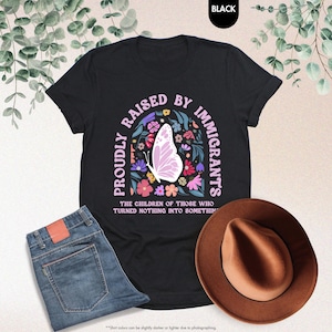 May include: Black t-shirt with the text "PROUDLY RAISED BY IMMIGRANTS" in a floral design with a butterfly. The shirt is paired with blue jeans and a brown hat. The text "THE CHILDREN OF THOSE WHO TURNED NOTHING INTO SOMETHING" is also on the shirt.