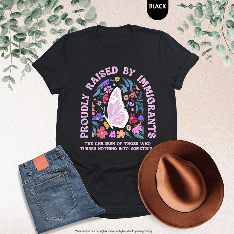 May include: Black t-shirt with the text "PROUDLY RAISED BY IMMIGRANTS" in a floral design with a butterfly. The shirt is paired with blue jeans and a brown hat. The text "THE CHILDREN OF THOSE WHO TURNED NOTHING INTO SOMETHING" is also on the shirt.