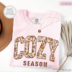 May include: A pale pink t-shirt featuring the words "COZY SEASON" in a brown, floral-patterned font. The shirt has a crew neck and short sleeves. A delicate necklace with a small pendant rests on the shirt. The words "MORE COLOR OPTIONS" are in a circle on the left.