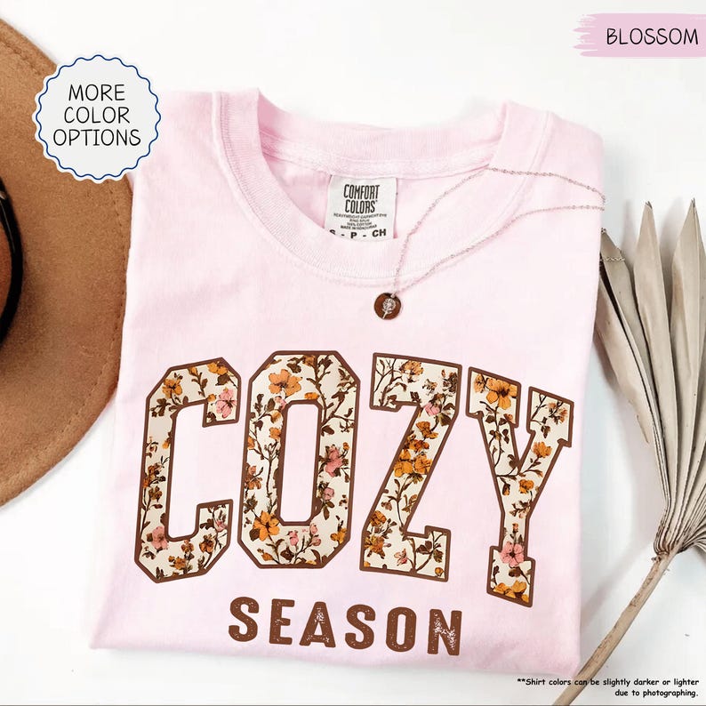 May include: A pale pink t-shirt featuring the words "COZY SEASON" in a brown, floral-patterned font. The shirt has a crew neck and short sleeves. A delicate necklace with a small pendant rests on the shirt. The words "MORE COLOR OPTIONS" are in a circle on the left.