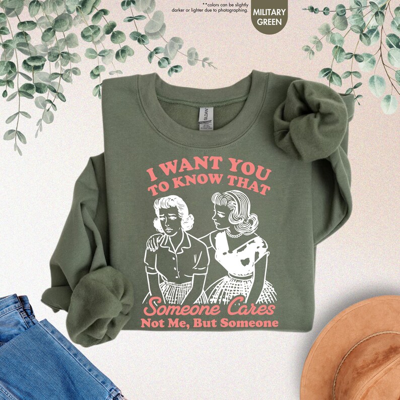 May include: Military green sweatshirt with the text "I WANT YOU TO KNOW THAT Someone Cares Not Me, But Someone" in pink and a graphic of two women. The long-sleeved top is folded, with a pair of blue jeans and a hat in the background.