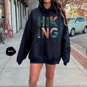 Pode incluir: Camisola de moletom com capuz preta com uma estampa gr&aacute;fica da palavra "HIKING" com cenas da natureza incorporadas nas letras.