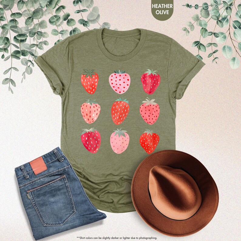 May include: A green t-shirt with a graphic of nine pink and red strawberries. The text "HEATHER OLIVE" is printed in white at the top of the shirt.