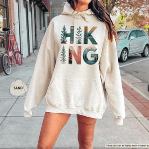 Pode incluir: Um moletom com capuz cor creme com uma estampa gr&aacute;fica da palavra "HIKING" em letras coloridas com designs inspirados na natureza. As letras s&atilde;o cercadas por imagens de montanhas, &aacute;rvores e plantas.