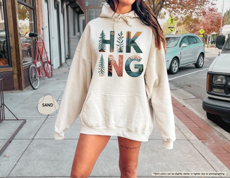 Pode incluir: Um moletom com capuz cor creme com uma estampa gr&aacute;fica da palavra "HIKING" em letras coloridas com designs inspirados na natureza. As letras s&atilde;o cercadas por imagens de montanhas, &aacute;rvores e plantas.