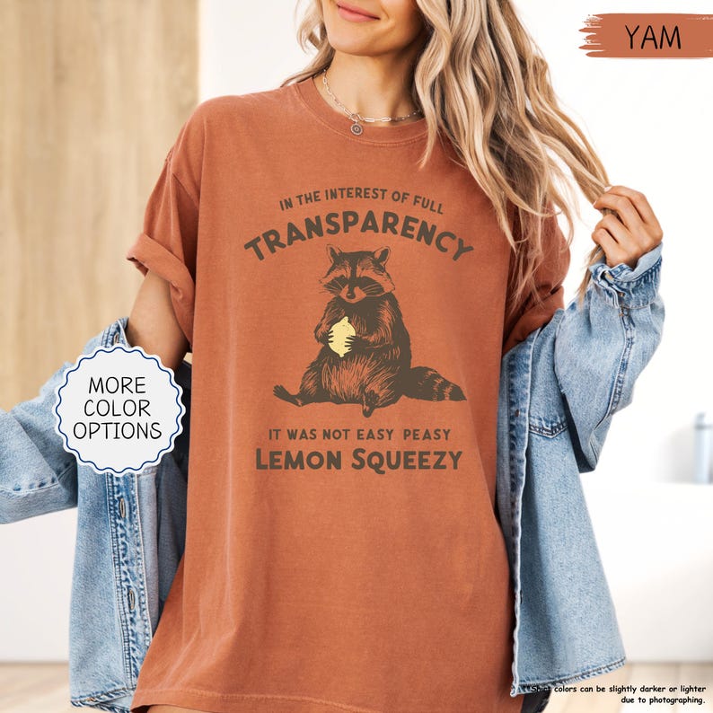 May include: A yam-colored t-shirt with a raccoon graphic holding a lemon. The text reads "IN THE INTEREST OF FULL TRANSPARENCY, IT WAS NOT EASY PEASY LEMON SQUEEZY". The shirt has rolled-up sleeves and a crew neck.
