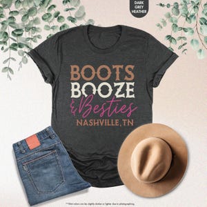 Pu&ograve; includere: T-shirt grigio scuro con la scritta "BOOTS BOOZE & Besties NASHVILLE, TN". L'immagine include anche un paio di jeans blu e un cappello color cammello.