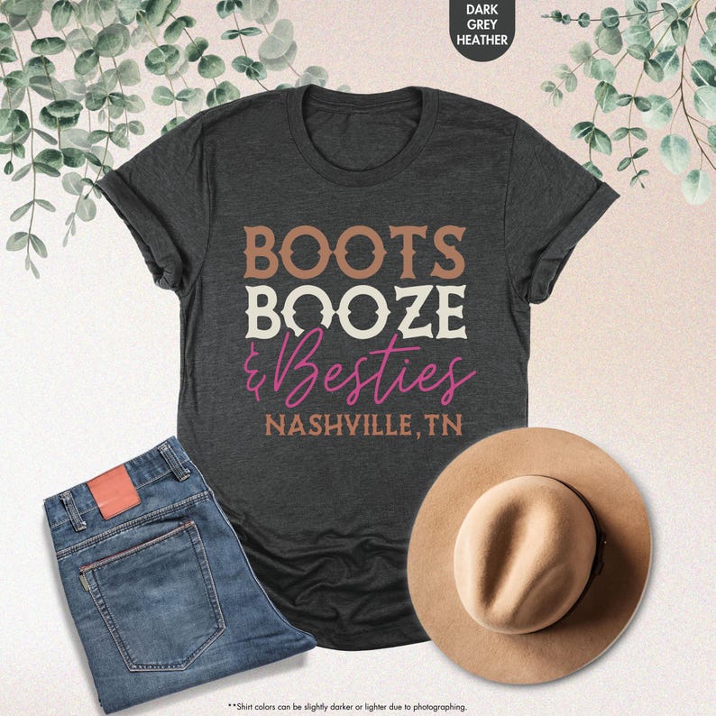 Pu&ograve; includere: T-shirt grigio scuro con la scritta "BOOTS BOOZE & Besties NASHVILLE, TN". L'immagine include anche un paio di jeans blu e un cappello color cammello.