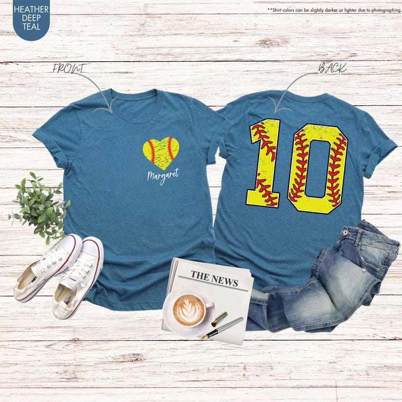 K&ouml;nnte beinhalten: Zwei T-Shirts in der Farbe Heide-Tiefblau mit einem Softball-Design. Das vordere T-Shirt hat einen herzf&ouml;rmigen Softball mit dem Namen "Margaret" darunter. Das hintere T-Shirt hat eine gro&szlig;e Nummer "10" aus einem Softball.