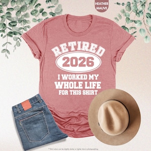 Puede incluir: Una camiseta color malva jaspeado con el texto "RETIRED 2026 I WORKED MY WHOLE LIFE FOR THIS SHIRT." La camiseta se combina con unos vaqueros azules y un sombrero fedora color canela. La imagen es una vista cenital.