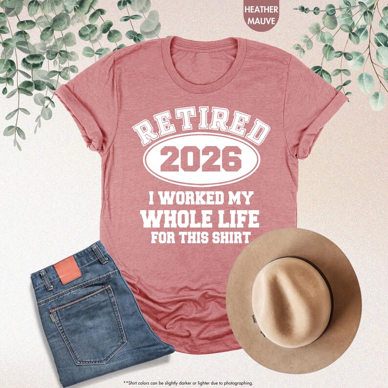 Puede incluir: Una camiseta color malva jaspeado con el texto "RETIRED 2026 I WORKED MY WHOLE LIFE FOR THIS SHIRT." La camiseta se combina con unos vaqueros azules y un sombrero fedora color canela. La imagen es una vista cenital.