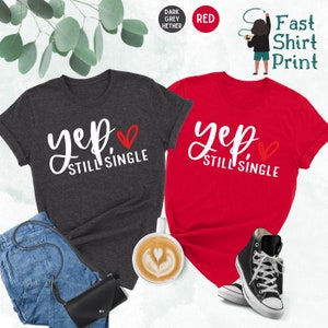 May include: Two t-shirts with the text "yep, still single" and a red heart. The shirts are in dark heather grey and red.
