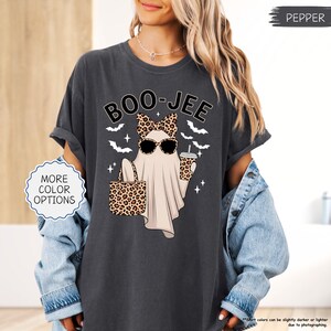 以下が含まれることがあります： ハロウィーンをテーマにしたグラフィックがプリントされた濃いグレーのTシャツ。サングラスとリボンをつけたお化けが、バッグとカップを持っています。「BOO-JEE」の文字がお化けの上にあります。Tシャツには「MORE COLOR OPTIONS」の文字も入っています。