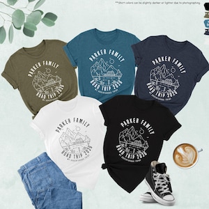 May include: Five t-shirts in various colours, including black, white, and olive green, are displayed. Each shirt features a circular graphic with the text "PARKER FAMILY ROAD TRIP 2026" and a mountain and lake design. A pair of jeans, a cup of coffee, and a pair of black trainers are also visible.