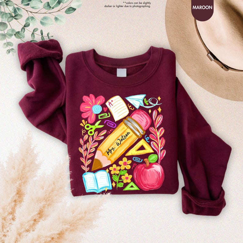 May include: Maroon sweatshirt with a colorful teacher-themed graphic. The design includes a large pencil with "Mrs. Watson" printed on it, an apple, flowers, scissors, and other school-related elements. The sweatshirt has long sleeves and a crew neck.