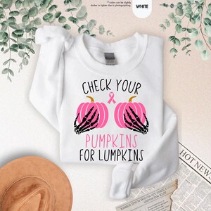 以下が含まれることがあります： 「CHECK YOUR PUMPKINS FOR LUMPKINS」の文字が入った白いスウェットシャツ。デザインは、黒い骸骨の手とピンクのリボンが付いた2つのピンクのかぼちゃが特徴です。クルーネックのスタイルのスウェットシャツです。