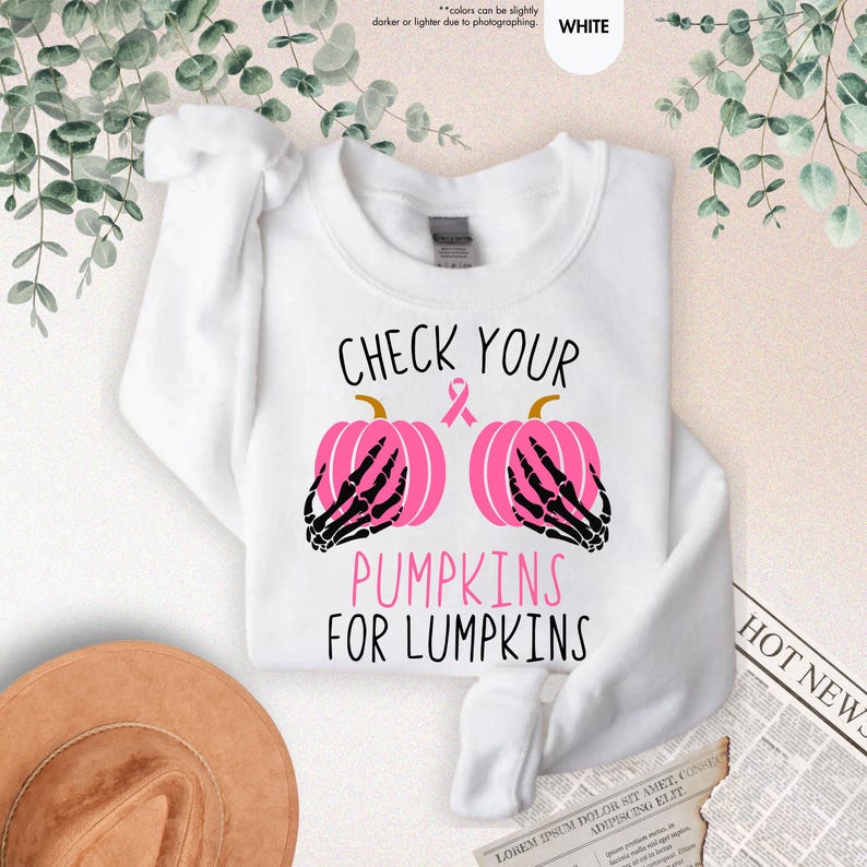 以下が含まれることがあります： 「CHECK YOUR PUMPKINS FOR LUMPKINS」の文字が入った白いスウェットシャツ。デザインは、黒い骸骨の手とピンクのリボンが付いた2つのピンクのかぼちゃが特徴です。クルーネックのスタイルのスウェットシャツです。