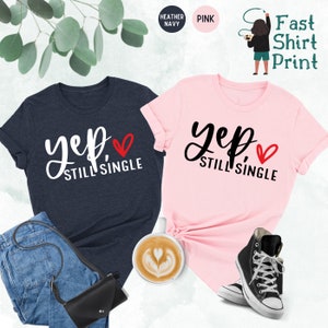 May include: Two t-shirts with the text "Yep, Still Single" and a red heart. The shirt on the left is heather navy and the shirt on the right is pink.
