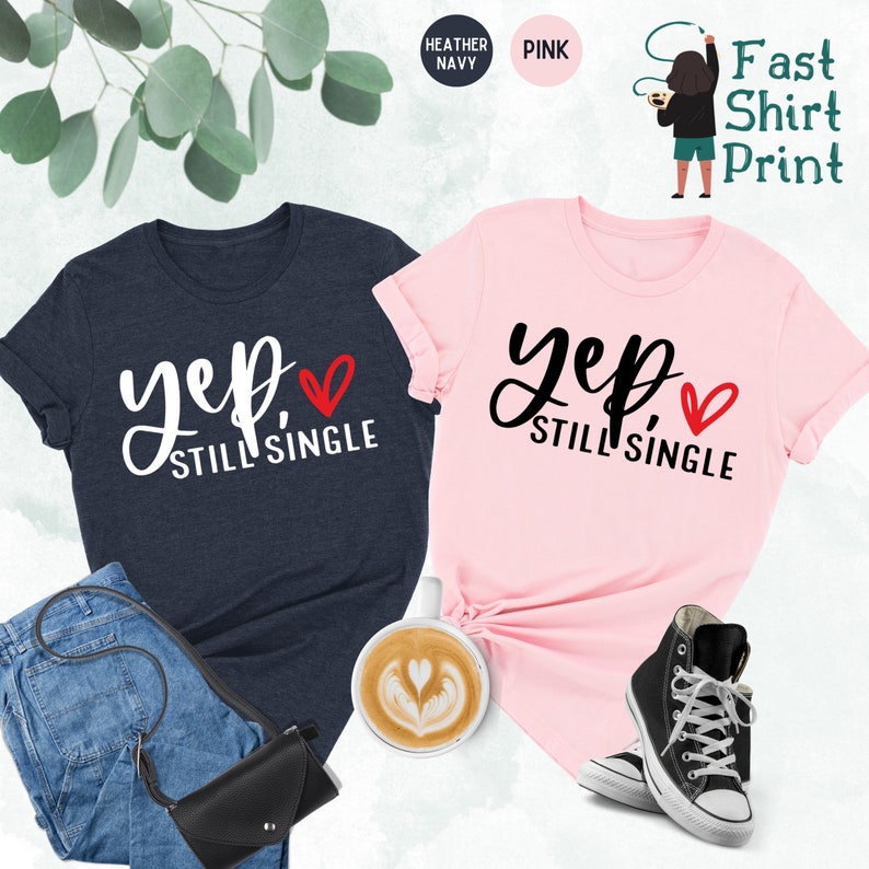 May include: Two t-shirts with the text "Yep, Still Single" and a red heart. The shirt on the left is heather navy and the shirt on the right is pink.
