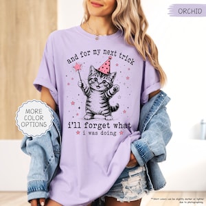 May include: An orchid-coloured t-shirt featuring a graphic of a tabby cat wearing a pink party hat and holding a star-tipped wand. The text reads, and for my next trick i'll forget what i was doing, surrounded by small pink stars and sparkles.