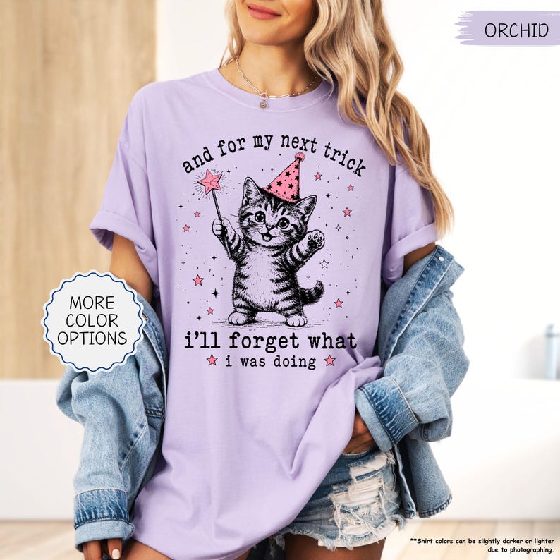 May include: An orchid-coloured t-shirt featuring a graphic of a tabby cat wearing a pink party hat and holding a star-tipped wand. The text reads, and for my next trick i'll forget what i was doing, surrounded by small pink stars and sparkles.