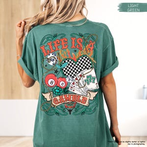 Puede incluir: Una camiseta verde claro con un estampado gr&aacute;fico de un coraz&oacute;n con un patr&oacute;n a cuadros, cartas de juego, cerezas y dados. El texto "Life is a Gamble" est&aacute; impreso en la camiseta.