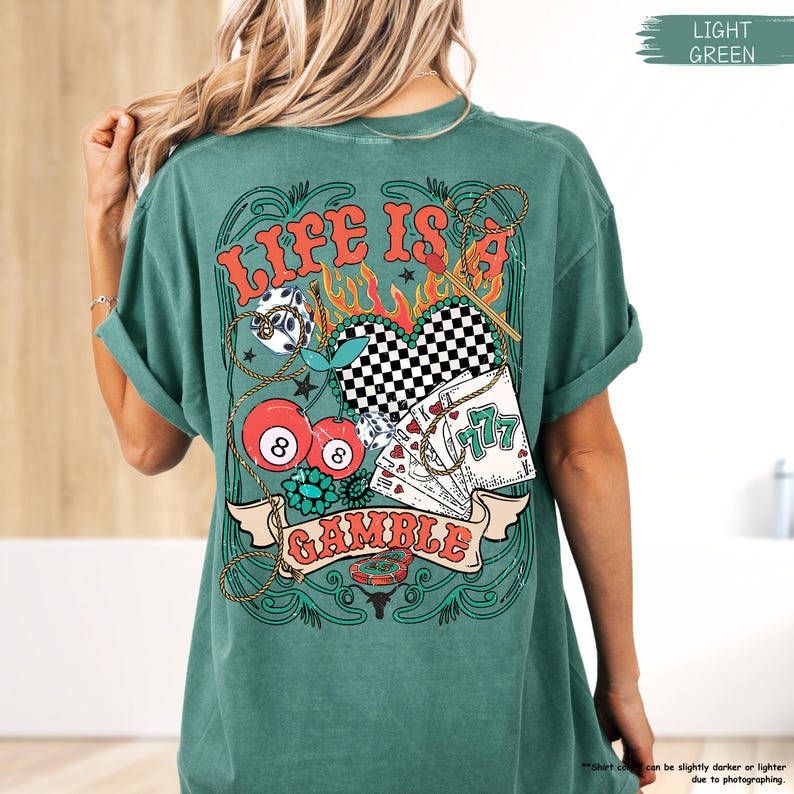 Puede incluir: Una camiseta verde claro con un estampado gr&aacute;fico de un coraz&oacute;n con un patr&oacute;n a cuadros, cartas de juego, cerezas y dados. El texto "Life is a Gamble" est&aacute; impreso en la camiseta.