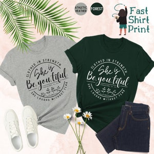 May include: Two t-shirts, one gray and one forest green, with the text "Clothed in Strength She is Beautiful" printed in a circular design. White sneakers and a pair of dark wash jeans are also visible. The phrase "She Laughs Without Fear" is also printed on the shirts.