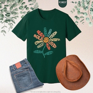 Pode incluir: Uma t-shirt verde escuro com um design floral feito de palavras como "corajoso" e "amado". A imagem tamb&eacute;m mostra cal&ccedil;as de ganga azuis e um chap&eacute;u de cowboy castanho. A palavra "FOREST" est&aacute; no topo.