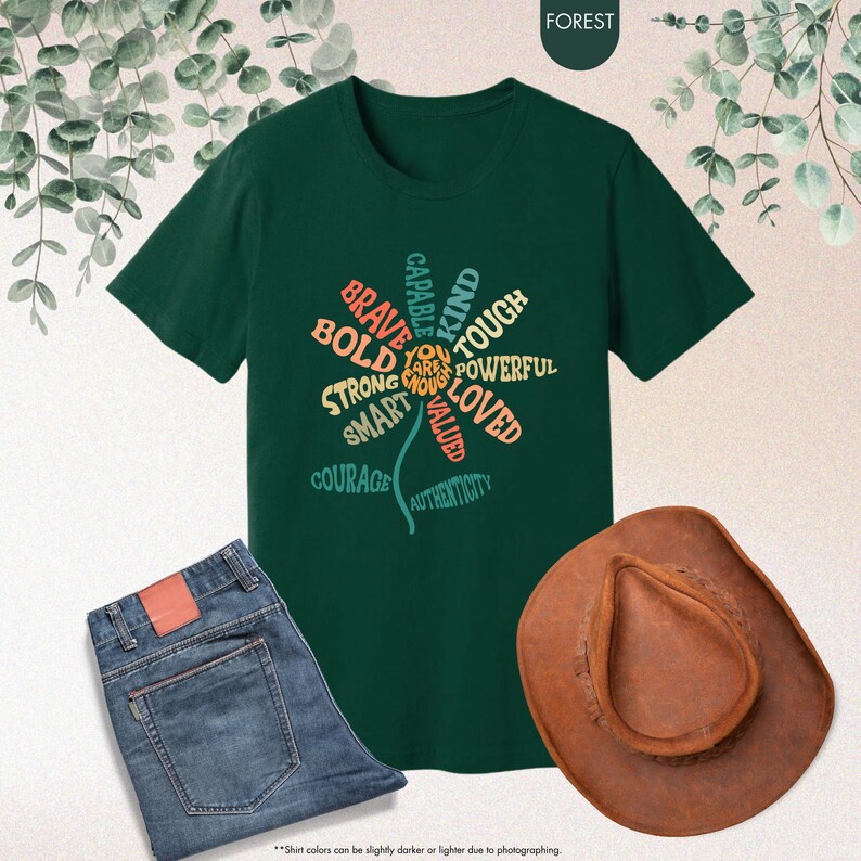 Pode incluir: Uma t-shirt verde escuro com um design floral feito de palavras como "corajoso" e "amado". A imagem tamb&eacute;m mostra cal&ccedil;as de ganga azuis e um chap&eacute;u de cowboy castanho. A palavra "FOREST" est&aacute; no topo.