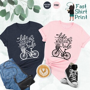 May include: Two t-shirts with a bicycle graphic and the text "My life is a beautiful ride". The t-shirts are in heather navy and pink.