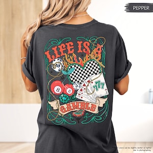 Puede incluir: Una camiseta gris oscuro con un estampado gr&aacute;fico de un coraz&oacute;n, cerezas, dados, cartas de juego y una cuerda. El gr&aacute;fico est&aacute; rodeado por un dise&ntilde;o verde y dorado. El texto "LIFE IS A GAMBLE" est&aacute; impreso en la camiseta.