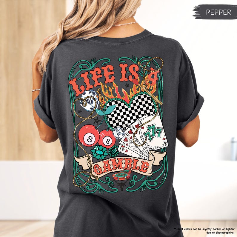Puede incluir: Una camiseta gris oscuro con un estampado gr&aacute;fico de un coraz&oacute;n, cerezas, dados, cartas de juego y una cuerda. El gr&aacute;fico est&aacute; rodeado por un dise&ntilde;o verde y dorado. El texto "LIFE IS A GAMBLE" est&aacute; impreso en la camiseta.