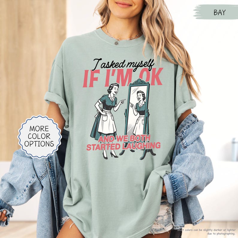 May include: A light green t-shirt with the text "I asked myself IF I'M OK and we both started laughing." The graphic features a vintage illustration of a woman looking in a mirror. The shirt also has a "More Color Options" button.