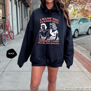 May include: Black hoodie with the text "I WANT YOU TO KNOW THAT Someone Cares Not Me, But Someone" in a retro font. The graphic features two women in vintage attire. The hoodie is a casual, oversized style.