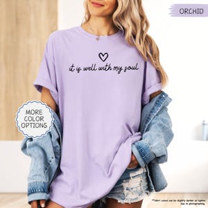 K&ouml;nnte beinhalten: Orchideenfarbenes T-Shirt mit dem Schriftzug "it is well with my soul" und einem Herz. Das Shirt besteht aus einem weichen, bequemen Material. Das Bild zeigt auch eine Jeansjacke und Shorts.