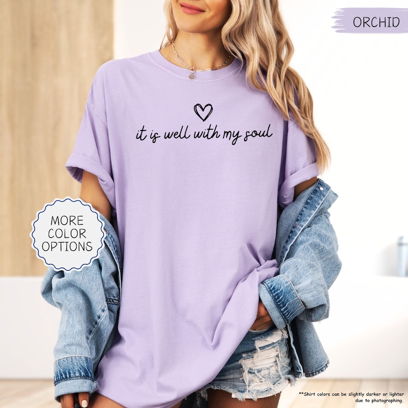 K&ouml;nnte beinhalten: Orchideenfarbenes T-Shirt mit dem Schriftzug "it is well with my soul" und einem Herz. Das Shirt besteht aus einem weichen, bequemen Material. Das Bild zeigt auch eine Jeansjacke und Shorts.