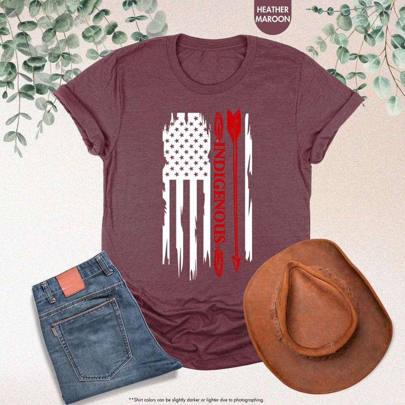 May include: A heather maroon t-shirt with a white distressed American flag design and the word "INDIGENOUS" in red lettering. Two red arrows are on the right side of the flag.