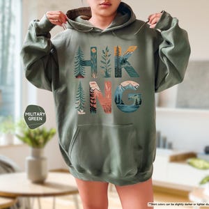 Pode incluir: Uma camisa de moletom verde com capuz com a palavra "HIKING" impressa na frente. As letras s&atilde;o compostas por ilustra&ccedil;&otilde;es de montanhas, &aacute;rvores e outras cenas da natureza.