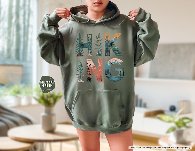 Pode incluir: Uma camisa de moletom verde com capuz com a palavra "HIKING" impressa na frente. As letras s&atilde;o compostas por ilustra&ccedil;&otilde;es de montanhas, &aacute;rvores e outras cenas da natureza.