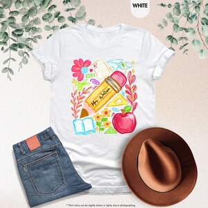 May include: White t-shirt with a colorful teacher-themed graphic. The design includes a large pencil with "Mrs. Watson" written on it, an apple, flowers, and school supplies. A pair of blue jeans and a brown hat are also in the image.