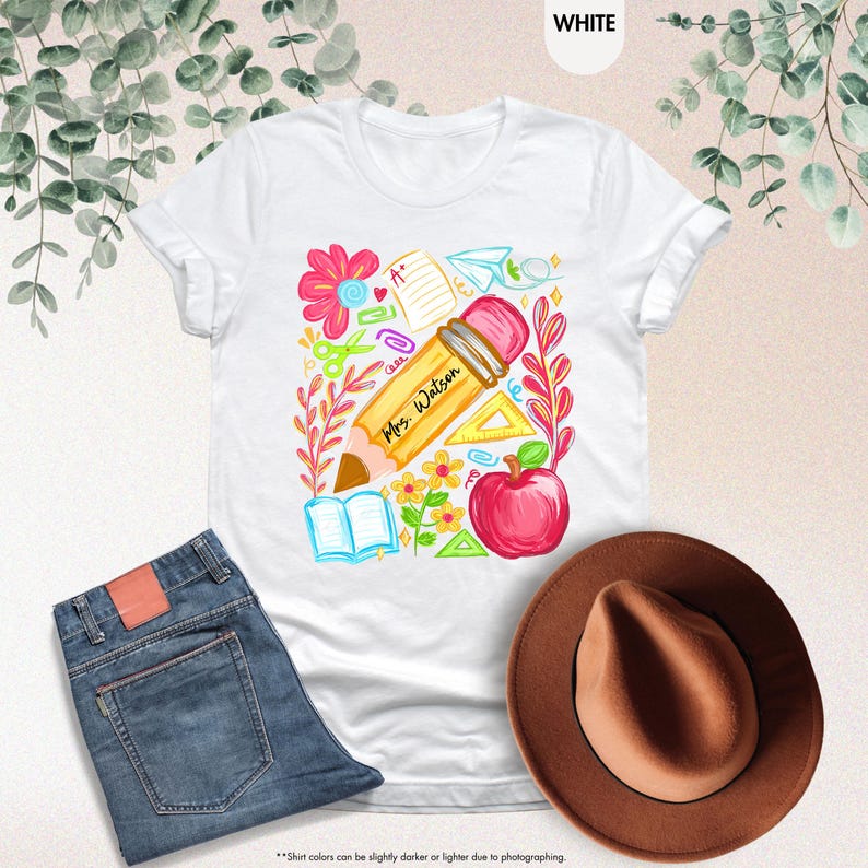 May include: White t-shirt with a colorful teacher-themed graphic. The design includes a large pencil with "Mrs. Watson" written on it, an apple, flowers, and school supplies. A pair of blue jeans and a brown hat are also in the image.