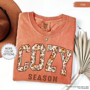May include: A yam-coloured sweatshirt featuring the words "COZY SEASON" in a floral design. The shirt has a crew neck and a small tag that says "COMFORT COLORS". A gold necklace with a pendant is draped across the shirt.