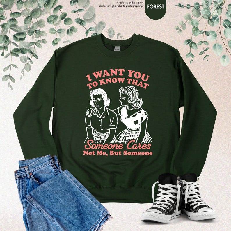 May include: A dark green sweatshirt with the text "I WANT YOU TO KNOW THAT Someone Cares Not Me, But Someone" in pink. The graphic features a vintage illustration of two women. Blue jeans and black sneakers are also in the image.