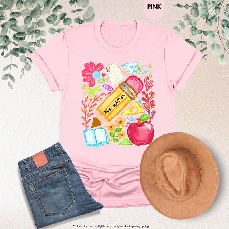 May include: Pink t-shirt with a colorful school-themed graphic. The design includes a large pencil with "Mrs. Watson" written on it, an apple, flowers, and school supplies. A pair of blue jeans and a brown hat are also in the image.