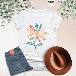 Pode incluir: Camiseta branca com um design floral composto por palavras positivas como "Brave", "Kind" e "Loved". A camiseta &eacute; combinada com jeans azuis e um chap&eacute;u fedora marrom. O texto "YOU ARE ENOUGH" est&aacute; no centro da flor.