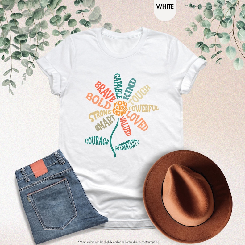 Pode incluir: Camiseta branca com um design floral composto por palavras positivas como "Brave", "Kind" e "Loved". A camiseta &eacute; combinada com jeans azuis e um chap&eacute;u fedora marrom. O texto "YOU ARE ENOUGH" est&aacute; no centro da flor.