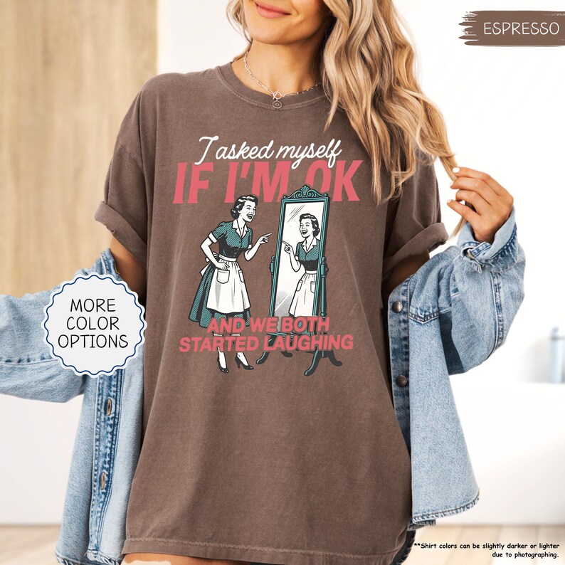 May include: Espresso-colored t-shirt with the text "I asked myself IF I'M OK" and "AND WE BOTH STARTED LAUGHING". The graphic features a vintage illustration of a woman looking in a mirror. The shirt has rolled sleeves and is paired with a denim jacket.